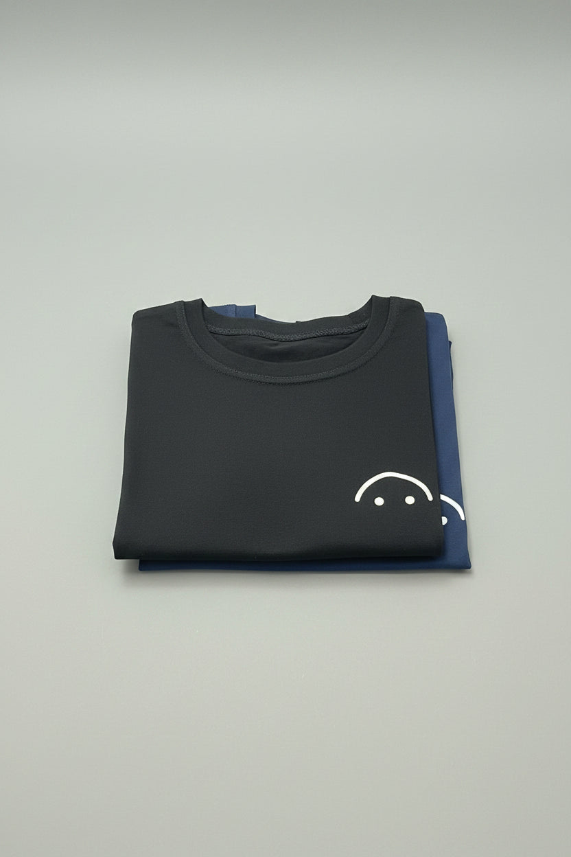 Two folded short-sleeve t-shirts on a wooden table, with the front shirt in black featuring a small white sad-face graphic and the back shirt in navy blue.