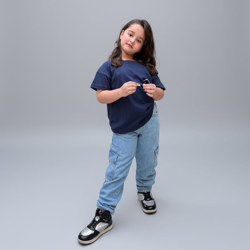 A young girl stands on a tiled patio wearing a navy blue t-shirt, light blue jeans, and black and white sneakers, surrounded by large potted plants.