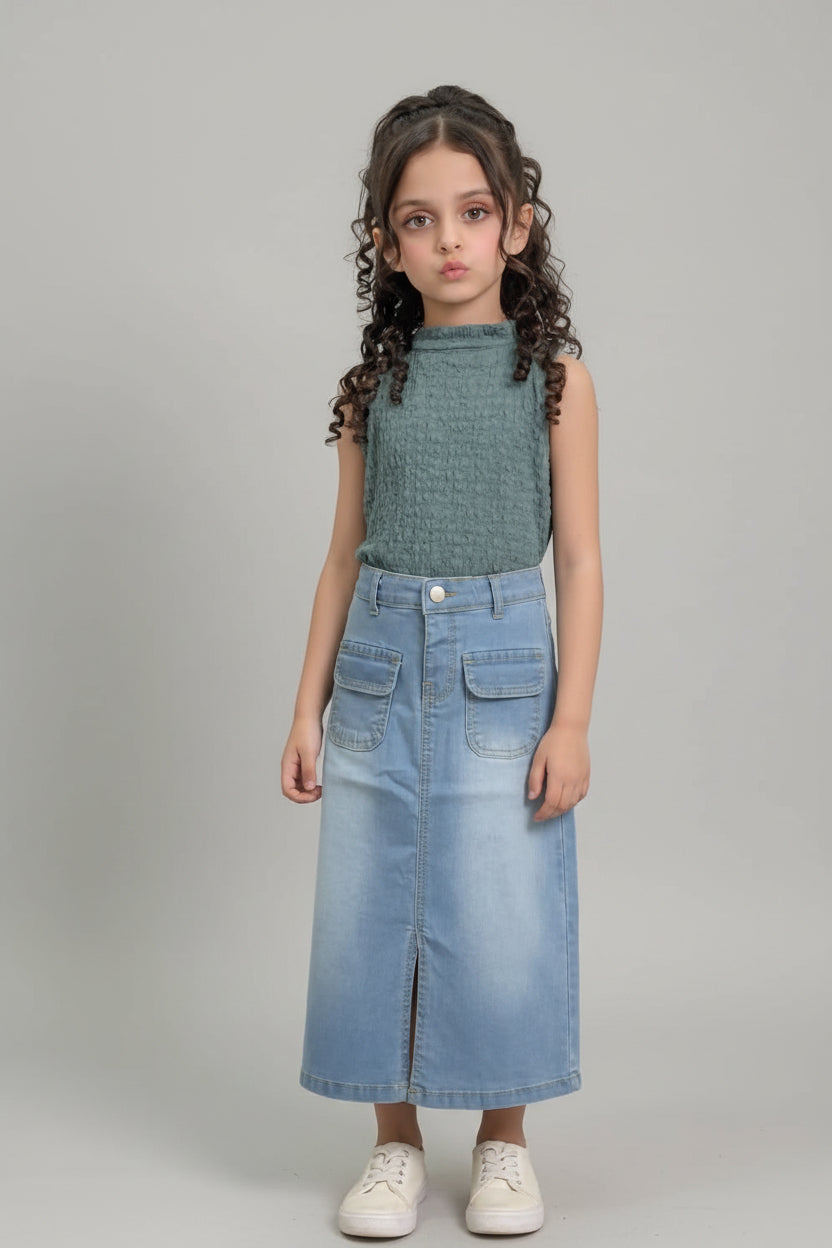 A young girl with curly brown hair stands indoors modeling a sleeveless textured sage green top and a light blue denim midi skirt with front patch pockets, paired with white sneakers.