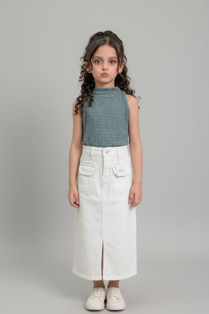 A young girl with curly brown hair stands indoors modeling a sleeveless textured sage green top and a long white denim skirt with front pockets and a side slit. She is wearing white sneakers.