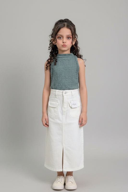A young girl with curly brown hair stands indoors modeling a sleeveless textured sage green top and a long white denim skirt with front pockets and a side slit. She is wearing white sneakers.