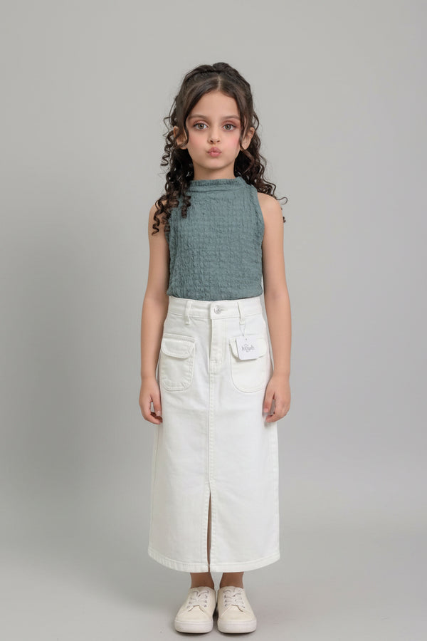 A young girl with curly brown hair stands indoors modeling a sleeveless textured sage green top and a long white denim skirt with front pockets and a side slit. She is wearing white sneakers.