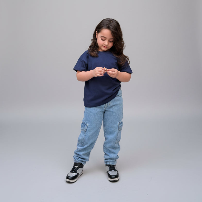 A young girl with long brown hair stands on a tiled patio wearing a navy blue t-shirt, light wash blue jeans, and black and white sneakers. She is surrounded by large potted green plants against a dark brick wall.
