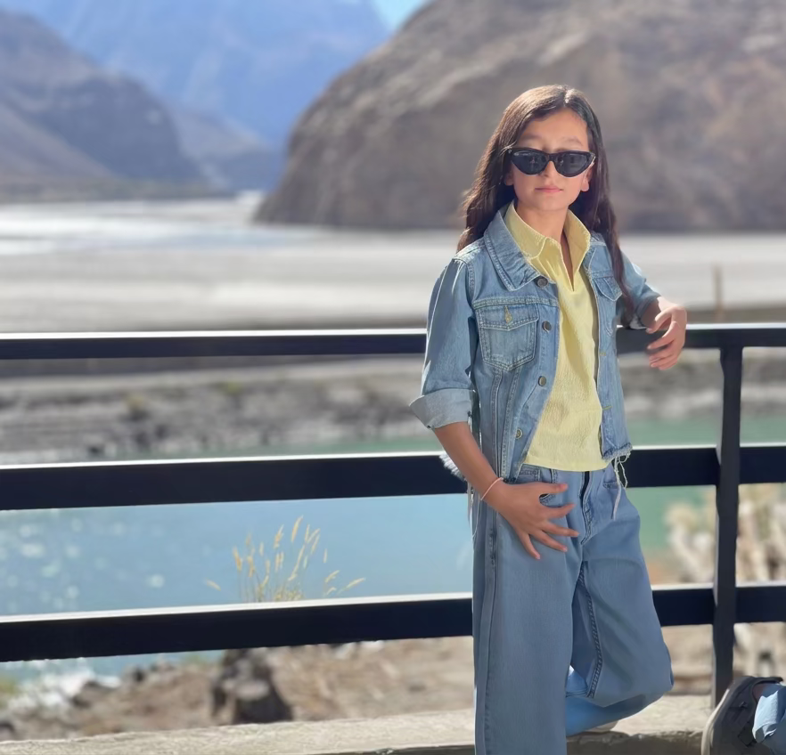 A young girl wearing a light blue denim jacket, matching wide-leg jeans with small white speckles, and a pale yellow polo shirt, posing outdoors on a balcony with a mountain and lake in the background.