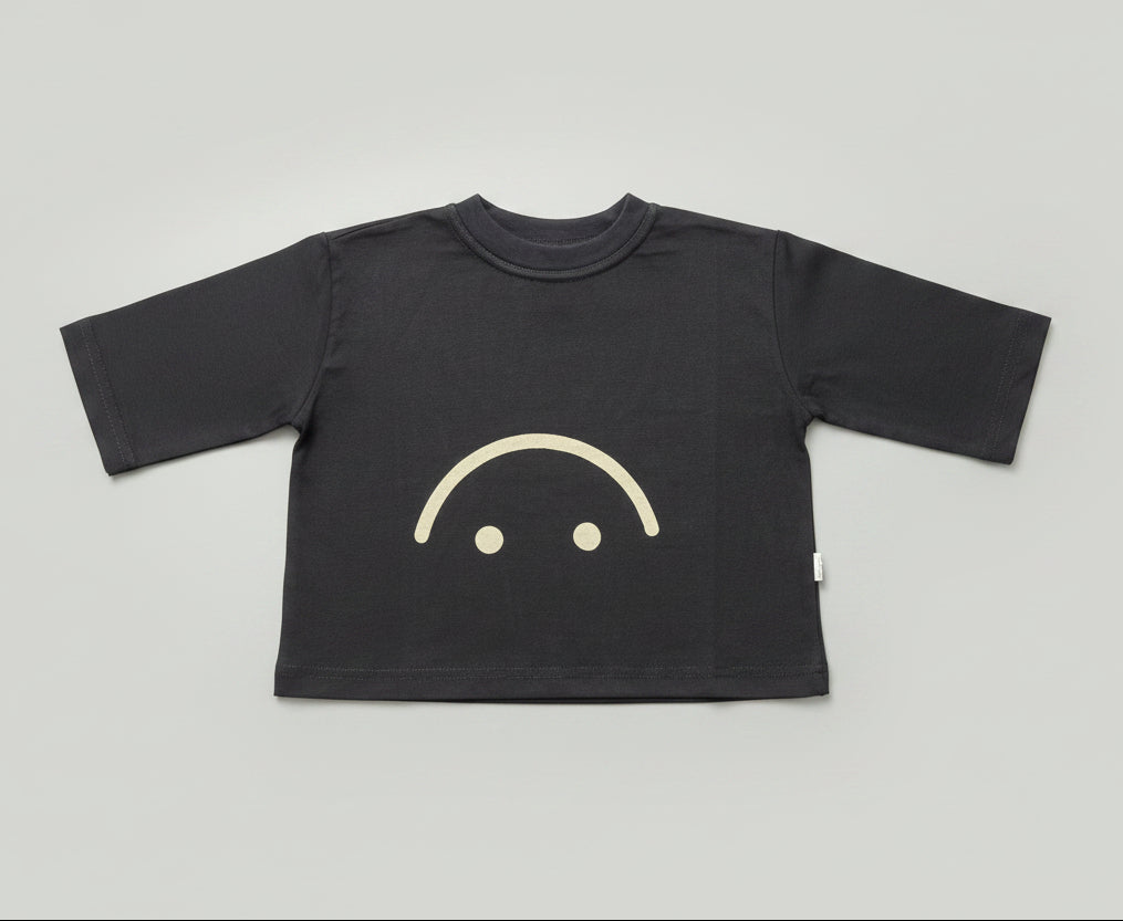 A black long-sleeve t-shirt with a simple white smiley face graphic on the front, laid flat on a wooden floor.