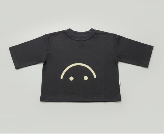 A black long-sleeve t-shirt with a simple white smiley face graphic on the front, laid flat on a wooden floor.