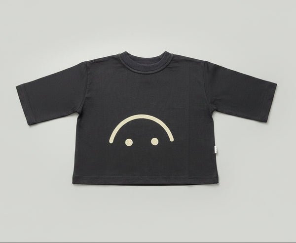 A black long-sleeve t-shirt with a simple white smiley face graphic on the front, laid flat on a wooden floor.