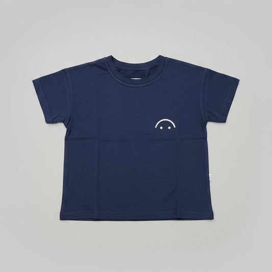 A navy blue short-sleeve crewneck t-shirt with a small white smiley face graphic on the left chest, laid flat on a wooden table.