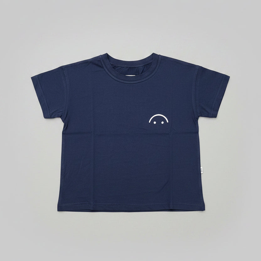 A navy blue short-sleeve crewneck t-shirt with a small white smiley face graphic on the left chest, laid flat on a wooden table.