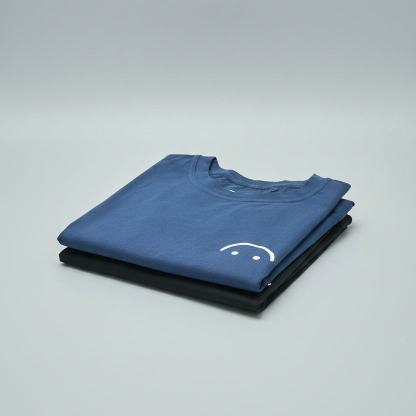 A folded blue t-shirt with a small white graphic on the chest, layered on top of a folded black t-shirt, both placed on a wooden surface.