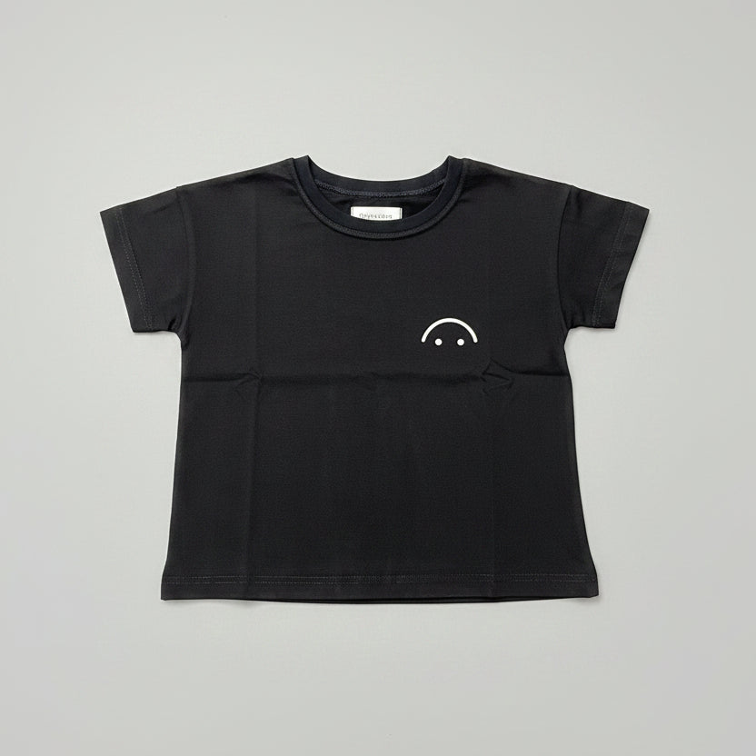 A black short-sleeve crewneck t-shirt with a small white smiley face graphic on the left chest, laid flat on a wooden table.