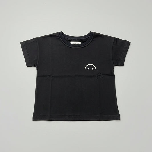A black short-sleeve crewneck t-shirt with a small white smiley face graphic on the left chest, laid flat on a wooden table.
