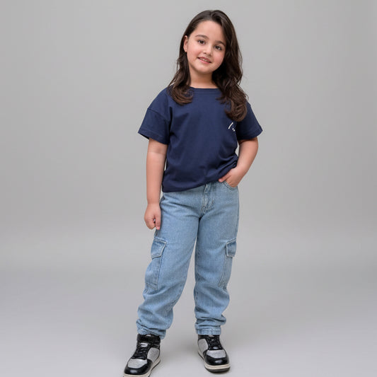 A young girl stands in a tiled courtyard wearing a navy blue t-shirt, light blue cargo jeans, and black and white sneakers, surrounded by large potted plants.