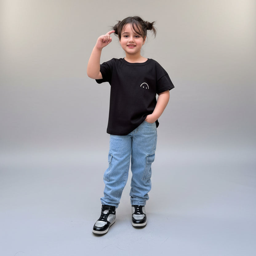 A young girl with her hair in two buns stands on a tiled patio wearing a black t-shirt with a small white graphic, light blue jeans, and black sneakers. She is posing in front of large potted plants and a glass door.